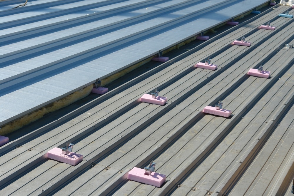 Standing seam metal roof with attachment clips and pink insulation spacers prepared for new roof panel installation.